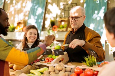 Çok ırklı müşteriler çiftçi pazarından alışveriş yaparken taze organik meyve tadıyorlar. Dost canlısı yaşlı çiftlik sahibi, tüketicilere satın almadan önce sezonluk gıda ürünlerini denemelerini teklif ediyor..