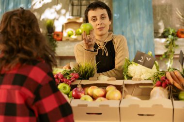 Çiftçi pazarında kameraya bakarken elinde yeşil elma tutan kadın satıcının portresi. Kadın tüccar tezgahın arkasında duruyor ve taze toplanmış meyve ve sebze satıyor..