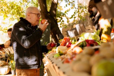 An elderly client smells bio apples and stands near farmers food market stand filled with fresh pesticide-free fruits and vegetables. Senior old man purchases organic produce grown locally. clipart