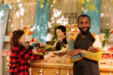 African american guy providing freshly harvested organic produce at greenmarket booth. Young small farm business owner standing near marketplace booth, grasping natural bio vegetable. clipart