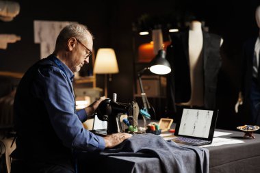 Yaşlı bavulcu atölye atölyesinde endüstriyel dikiş makinesiyle tekstil kumaşı kesiyor. Tecrübeli eski terziler, müşteri temsilciliği için enfes kıyafetler üretiyor.