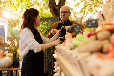 Elderly man and young seller arranging various freshly harvested produce on greenmarket stand. Local merchants talking while arranging eco friendly fruits and vegetables at farmers market booth. clipart