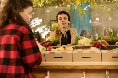 Neşeli kadın, elinde elma tutarken sağlıklı organik ürünler satarak market standının arkasında duruyor. Kadın manav kameraya bakarken taze yetişmiş meyve ve sebzeler sergiliyor..