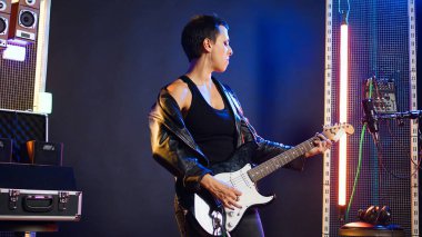 Baş parmaklarını gösteren ve gitar çalan bir kadın, stüdyoda müzik performansı. Kadın rockçı, olumlu bir işaretle onayını veriyor. Kamera A.