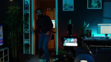 İçerik üreticisi ev stüdyosuna giriyor, canlı yayında kameraya doğru serin el hareketleriyle hareket ediyor. Gösterici işaret ediyor, büyüleyici seyirci, kamera A