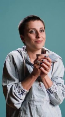 Dikey video Mutlu kadın mavi arka planda bir fincan kahve yudumluyor, stüdyoda sıcak kafein içeceklerinin keyfini çıkarıyor. Modern kıyafetli, kendine güvenen ve içecek içen biri. Kamera A.