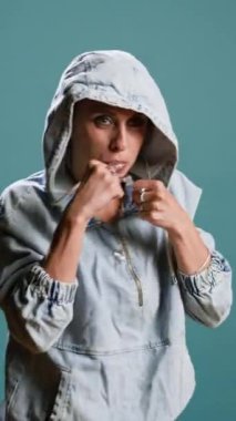 Dikey video güçlü cesur kadın kaslarını kasıyor ve stüdyoda yumruklar savuruyor, mavi arka planda sokak dövüşü kavramı. Saldırgan, kendine güvenen ve dövüşmeye hazır biri. Kamera A.