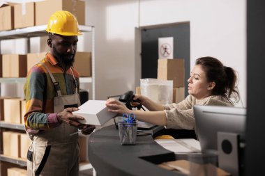 Depo amiri, beyaz karton kutuyu tarayıcı kullanarak tarıyor, Afrikalı Amerikalı bir çalışanla nakliye bölümlerini tartışıyor. Çeşitli ekipler depoda kalite kontrolde çalışıyor.