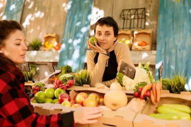 Kadın müşteri meyve ve sebze seçerken, satıcı da elinde elmayla tezgahın arkasında duruyor. Hasat fuarında çeşitli mevsimsel organik ürünler satan kadın çiftçi portresi.