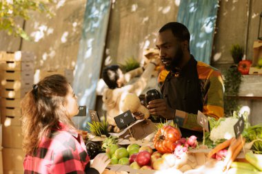 Zenci adam çiftçi pazarından doğal sağlıklı ürünler gösteriyor, yerel olarak yetişmiş meyve ve sebzeler. Erkek çiftçi, çeşitli mevsimsel organik sebze ürünleri hakkında müşteriyle konuşuyor..