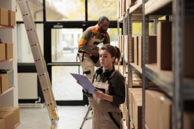 Küçük işletme çalışanı elinde pano tutarak listeyi analiz ediyor, depoda nakliye için ürünler hazırlamaya başlamadan önce müşterilerin siparişlerine bakıyor. Personel depoda koruyucu giysi giyiyor.