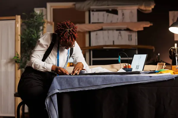 stock image Expert focused suitmaker creating bespoken sartorial fashion collection surrounded by sewing materials and sketch drawings. African american couturier working in exquisite tailoring studio.