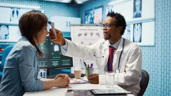 stock image African american specialist consulting a young woman in medical office, discussing lab test results and other analysis for wellness. Medic takes her temperature with a digital thermometer. Camera B.