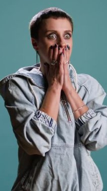 Dikey video, ürkek bir kadının mavi arka planda ağzını kapatarak korkunç bir şeye bakmasına neden oldu. Sokak tarzı olan biri endişeli ve korkmuş, endişeli bir kız.