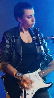 Dikey video bağımsız kadın elektro gitarını karanlık bir stüdyoda spot ışıklarıyla canlı performans için çalıyor. Deri ceketli, kendine güveni tam bir sanatçı.