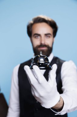 Porter with gloves holds service bell to show professional hotel concierge occupation, giving assistance to people. Young man employee in suit posing in studio, luxury hospitality industry. clipart