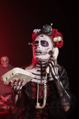 Meksika dios de los muertos bayramında telefona cevap vermek için ev telefonu kullanan ürkütücü kadın. La Catrina gibi giyinmiş biri, ölüm gününü kutluyor ve ofis kablosu ile konuşuyor.