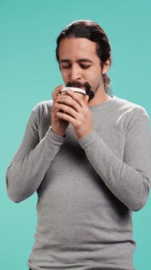 Dikey Videocu yorgun hissediyor, işe giderken uyanmak için kahve içiyor. Sabah enerji kazanmak için kafeinli içecek kullanan bitkin bir insan, mavi stüdyo arka planı, kamera B