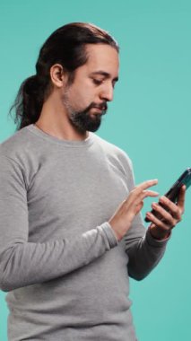 Akıllı telefon dokunmatik ekranında kayan gülümseyen bir adamın dikey video portresi, sosyal medya beslemelerini kontrol etmek için cep telefonu kullanıyor. Neşeli insan internet sitelerine bakıyor, stüdyo arka planında izole edilmiş.