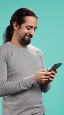 Dikey video Mutlu Adam akıllı telefon kullanarak en iyi arkadaşına mesaj atıyor, mesajları okuyor. Rahatlamış insan cep telefonuyla arkadaşlarıyla sohbet ederken eğleniyor, mavi stüdyo arka planında izole edilmiş, kamera B