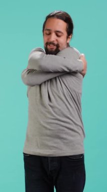 Stüdyoda duran bir adamın dikey video portresi, stüdyo arka planında izole edilmiş rahatlık ve kendini sevme jesti ile kendine sarılıyor. Neşeli insan kendini kabul ediyor, kendini kucaklıyor.