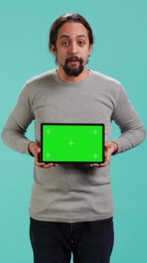 Yeşil ekran tableti sunan dikey video etkileyicisi, stüdyo arka planında izole edilmiş. Arap kişi, reklam markaları, stüdyo arkaplanı, kamera A için kullanılan boş krom anahtar cihazını elinde tutuyor.