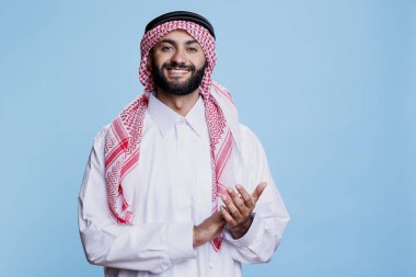 Gülümseyen Müslüman adam alkış hareketi yapıyor ve heyecanlı bir ifadeyle kameraya bakıyor. İslami tobe ve ghutra giyen neşeli Arap performans stüdyosu portresini alkışlıyor.