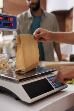 Kasiyer masasındaki bir poşette biyolojik gıda ürünlerinin ağırlığını kontrol eden bir çift beyaz erkek elinin detaylı görüntüsü. Vezne tezgahının üzerinde tartıyla ilgili kağıtları tutan kişiye yakın çekim.