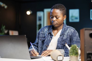 Evde çalışan, not defterine yazan siyahi bir kadın. Bir dizüstü bilgisayar kullanan, birden fazla iş yapan, not alan Afrikalı Amerikalı bir serbest yazar. Yoğunlaşmış ve verimli bir şekilde çalışmaya kararlı..