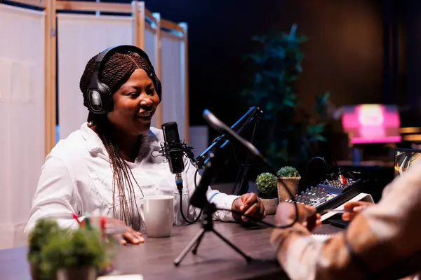 stock image Cheerful female african american influencer speaking into microphone, answering questions from radio host in home studio. Joyful black woman using recording equipment for online talk show.