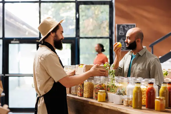 stock image Middle Eastern male customer smells yellow fruit, that looks like a lemon, while caucasian storekeeper wearing a hat and apron looking over to the other side of counter holding a package.