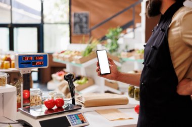 Depo görevlisi taze ürünleri tartarken aynı zamanda cep telefonunu çevre dostu bir markette boş bir model şablonuyla kavrıyor. Beyaz önlük takan ve elinde beyaz perdeli akıllı telefon tutan beyaz bir adam..