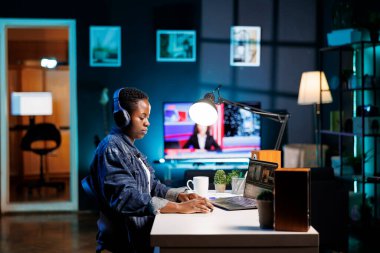 Afrikalı Amerikalı kadın kulaklık takarken video düzenlemek için kişisel bilgisayarını kullanıyor. Yaratıcı işlere dalmış, müzik dinleyen ve dizüstü bilgisayarını film görüntülerini incelemek için kullanan siyahi bir kadın..