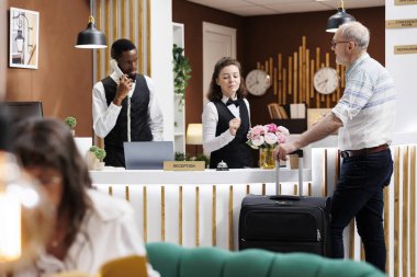 Otel resepsiyonunda, arkadaş canlısı personel emekli kıdemli adamı karşılıyor ve check-in yardımı sağlıyor. Lüks tatil beldesindeki en iyi tatil etkinliklerinin kapıcılarından tavsiye isteyen yaşlı erkek turist.