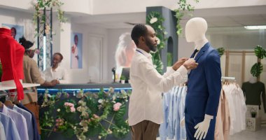 Lüks moda mağazasında çalışan titiz perakende asistanı blazer ceket cebinde cep mendili ayarlıyor. Deneyimli Afro-Amerikan çalışanı. Mankenler üzerinde yeni moda giysiler sergiliyor.