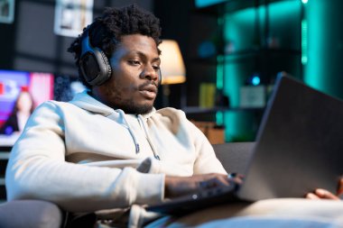 Afro-Amerikalı adam evde oturup kulaklık kullanarak dizileri izleyerek rahatlıyor. RGB 'de boş vakitlerin keyfini çıkaran kaygısız kişi gece oturma odasını aydınlatıyor ve eğlence medyasını tüketiyor.