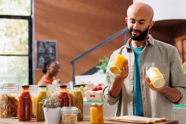 Middle Eastern man holds and analyzes two glass jars filled with natural honey in grocery store while black woman looks around. Male customer checking locally made organic food product. clipart