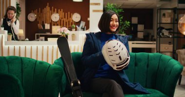 Otel lobisinde kış sporlarına hazırlanan kadın, Asyalı adam kabarık ceket ve koruyucu kaskla hazırlanıyor. Salon bölgesinde kayak ekipmanlarını kontrol eden turistler, mevsimsel aşırı hareketlilik.