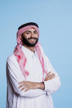 Geleneksel Müslüman başlığı ve cübbesi giyen mutlu Arap adam kendine güvenen poz portresi yapıyor. Beyaz mantolu gülümseyen İslami kişi kollarını kavuşturmuş kameraya bakıyor.
