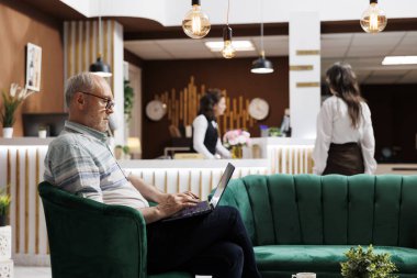 Yaşlı erkek turist göz gezdirirken ve dizüstü bilgisayarla oda ayırtırken otel resepsiyonuna kadın misafir giriş yapıyor. Emekli kıdemli adam kişisel bilgisayarını çevrimiçi kayıt için kullanıyor..