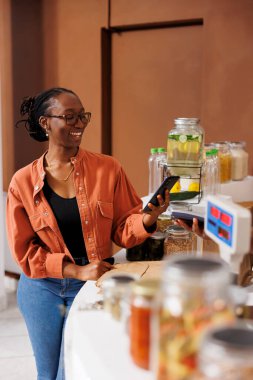 Cheerful african american customer using mobile device for cashless checkout, purchasing healthy bio food with nfc payment. Black woman holding cell phone and paying for her organic products. clipart