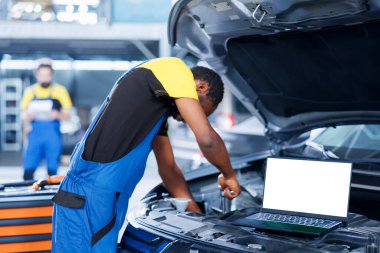 Kaputu açık arabadaki model dizüstü bilgisayar, bulanık arka plandaki tamirci motoru değiştirirken. Müşteri otomobilini tamir eden lisanslı garaj uzmanının yanındaki izole ekran cihazı