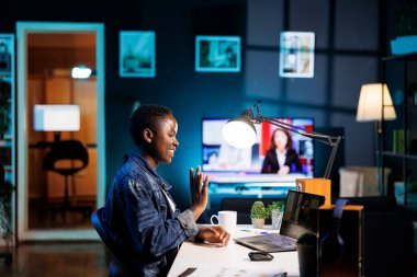 Afrikalı Amerikalı blogcu bir masada oturmuş video görüşmesi yaparken kişisel bilgisayarına el sallıyor. Siyahi kadın dijital teknolojiye dalmışken çevrimiçi sohbet ve araştırma için dijital dizüstü bilgisayar kullanıyor.
