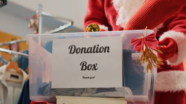 Close up shot of donation box in fashion shop set up for humanitarian help during Christmas holiday season. Worker dressed as Santa Claus collecting unneeded clothes from generous clients clipart