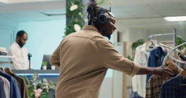 Süslü moda mağazasında dans eden Afrikalı Amerikalı bir adam şık kıyafetler ararken. Premium showroom 'dan tasarımcı kıyafetleri alırken kulaklıkla müzik dinleyen mutlu müşteri