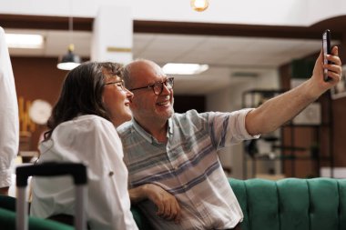 Yaşlı bir adam özel otel lobisinde eşiyle selfie çektirmek için cep telefonu tutuyor. Bavullu emekli yaşlı bir çift, ellerinde mobil cihazla oturma odasında oturuyorlar..