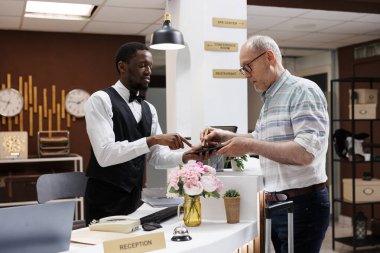 Otel resepsiyonunda duran olgun yaşlı bir adam. Kibar resepsiyonist dijital tablette rezervasyon prosedürüne yardım ediyor. Yaşlı erkek istemci akıllı aygıt üzerinde çevrimiçi bir kayıt formu imzalar.