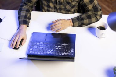 Erkek bireyin dizüstü bilgisayarına bakmak için fareyi kullanışının havadan görüntüsü. Elinde kahve fincanıyla masada oturan kişi evden çalışmak için modern teknolojiyi kullanıyor..