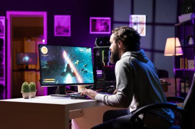 Man in dimly lit apartment playing singleplayer videogame with spaceship shooting laser streams at asteroid. Gamer using crosshair overlay to hit targets in science fiction arcade game clipart