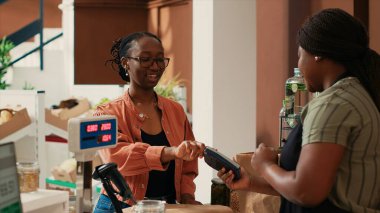 African american shopper paying with card at cash register, purchasing organic natural fruits and veggies for healthy nutrition. Smiling woman supporting local vendor and ecological farming. clipart